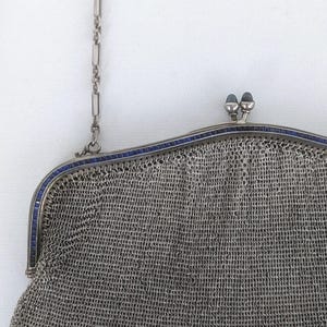 May include: A silver mesh purse with a blue enamel trim and a chain strap. The purse has a clasp closure with two blue enamel knobs.