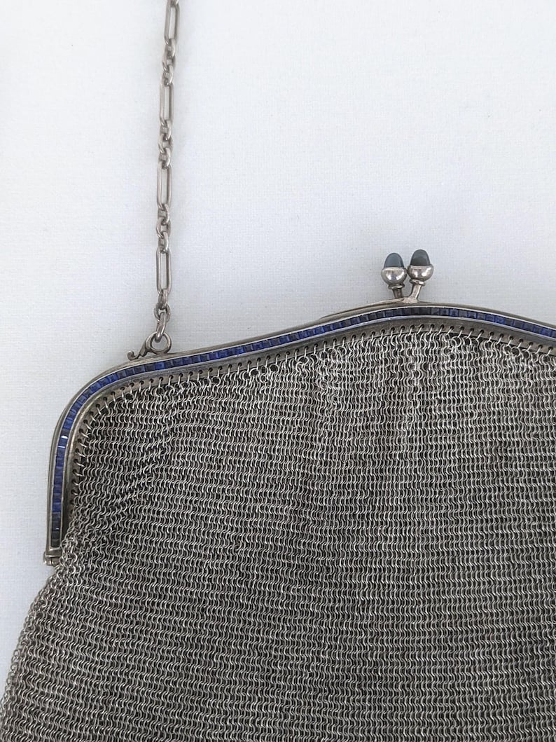 May include: A silver mesh purse with a blue enamel trim and a chain strap. The purse has a clasp closure with two blue enamel knobs.