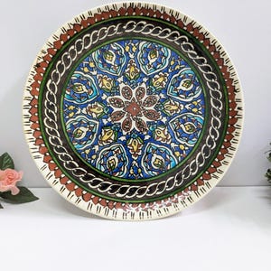 Puede incluir: Un plato decorativo redondo de cerámica con un diseño intrincado y colorido. El plato presenta un motivo floral central en tonos marrón, azul y amarillo, rodeado por un borde detallado de color verde, marrón y rojo. El borde exterior es de color crema.