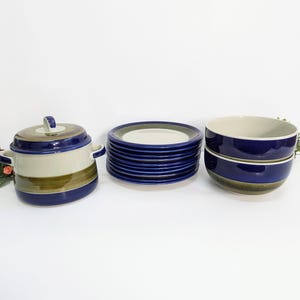 Può includere: Un set di stoviglie in ceramica vintage nei toni del blu, beige e verde oliva. Il set comprende una casseruola con coperchio, una pila di piatti e due ciotole. I pezzi presentano un design a righe, disposti su una superficie bianca.