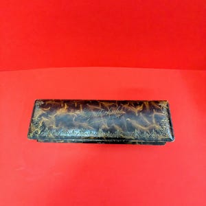 Puede incluir: Una caja rectangular antigua con un acabado oscuro y moteado y ribete dorado. La caja tiene un diseño ornamentado y texto en la tapa. El fondo es rojo liso.