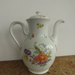 Può includere: Una caffettiera in porcellana bianca con un motivo floreale. La caffettiera ha un bordo dorato attorno al bordo e un coperchio con un pomo. I fiori sono in tonalità di rosa, arancio, giallo e viola.