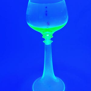 Puede incluir: Una copa de vino de cristal transparente con un tallo largo y texturizado y una base redonda. La copa está llena de un líquido verde brillante, sobre un fondo azul liso.