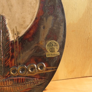 Op de afbeelding: Een ronde keramische wandhanger met een bruin, wit en gouden kleurenschema. De wandhanger heeft een gestileerd ontwerp van veren en cirkels. De wandhanger heeft een gouden label met de tekst "Ruscha Art echt Handwerk".