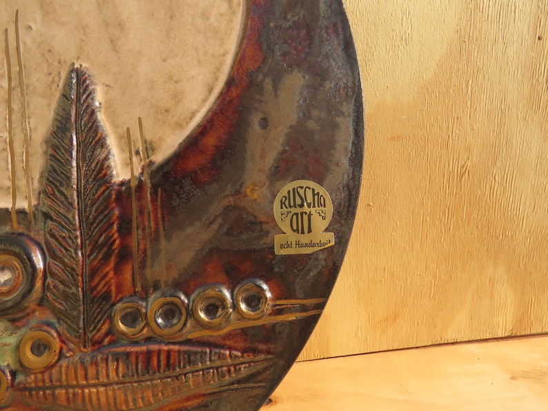 Op de afbeelding: Een ronde keramische wandhanger met een bruin, wit en gouden kleurenschema. De wandhanger heeft een gestileerd ontwerp van veren en cirkels. De wandhanger heeft een gouden label met de tekst "Ruscha Art echt Handwerk".