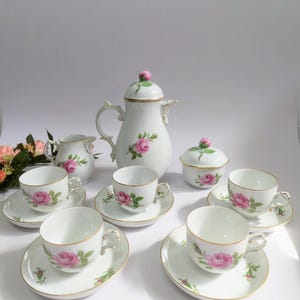 Può includere: Un servizio da tè in porcellana bianca con disegni floreali di rose rosa. Il set comprende una teiera, una zuccheriera, una lattiera e sei tazze da tè con piattini. Ogni pezzo ha finiture dorate.