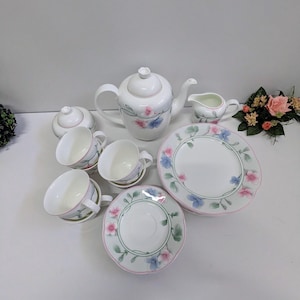 Könnte beinhalten: Ein weißes Porzellan-Teeservice mit floralen Designs. Das Set beinhaltet eine Teekanne, eine Zuckerdose, einen Sahnekännchen, Tassen, Untertassen und einen Teller. Das florale Muster zeigt rosa und blaue Blumen mit grünen Blättern.