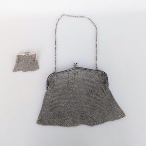 May include: A silver mesh purse with a chain strap. The purse is closed and has a scalloped edge. The chain strap is attached to the purse with two small silver clasps.