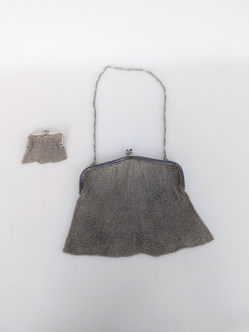 May include: A silver mesh purse with a chain strap. The purse is closed and has a scalloped edge. The chain strap is attached to the purse with two small silver clasps.