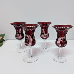 Pode incluir: Quatro taças de vinho vermelho rubi com desenhos gravados de flores e veados brancos. As taças têm um topo alargado e uma base redonda transparente. As taças são provavelmente para servir vinho ou outras bebidas.