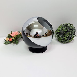 May include: A contemporary, spherical decorative item with a polished chrome finish and a black base. The sphere is divided by a diagonal line, with the top half in chrome and the bottom half in black. The object reflects the surrounding environment.
