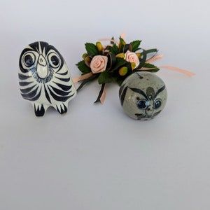 May include: Two ceramic owl figurines, one black and white with a striped pattern, the other grey with blue and white details. Both owls have large eyes and are sitting on a white surface. A small floral arrangement with pink roses and green leaves is in the background.