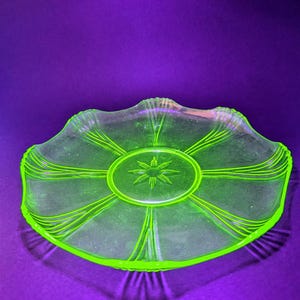 Puede incluir: Un plato de vidrio redondo, translúcido y de color verde neón, con un patrón central de estrella rodeado por un borde circular. El plato tiene un borde festoneado y líneas radiantes que se extienden desde el centro hacia el borde.