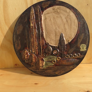 Op de afbeelding: Een ronde, keramische wandhanger met een textuur, bruin en gouden ontwerp. Het ontwerp heeft een grote, witte cirkel in het midden, omringd door gestileerde bomen en bladeren. De wandhanger is onderaan rechts gesigneerd met "Kunst im Off".