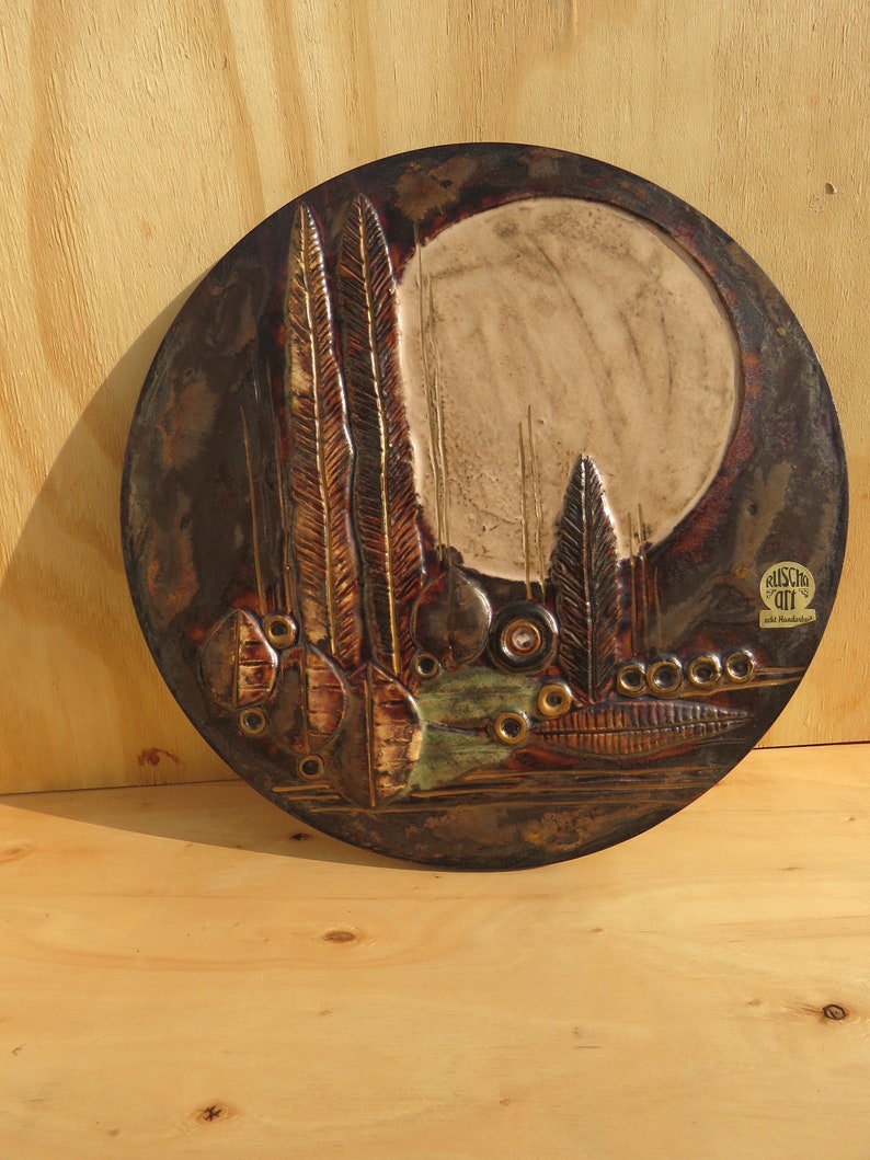 Op de afbeelding: Een ronde, keramische wandhanger met een textuur, bruin en gouden ontwerp. Het ontwerp heeft een grote, witte cirkel in het midden, omringd door gestileerde bomen en bladeren. De wandhanger is onderaan rechts gesigneerd met "Kunst im Off".
