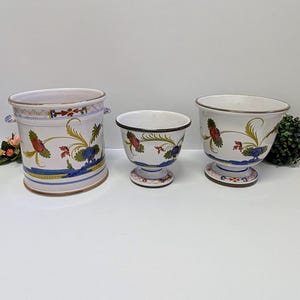 Könnte beinhalten: Drei weiße Keramik-Blumentöpfe mit floralen Designs. Die Töpfe variieren in Größe und Form, mit einem zylindrischen Topf links, einem Sockeltopf in der Mitte und einem schalenförmigen Topf rechts. Jeder hat rote, blaue und grüne Blumenmuster.