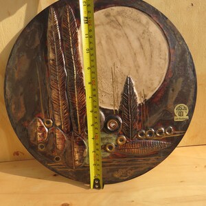 Op de afbeelding: Een rond keramisch wandkunstwerk met een bruine, roestkleurige en witte glazuur. De kunst toont een gestileerd ontwerp van bomen en een maan. Het stuk is ongeveer 38 cm in diameter. De kunst is rechtsonder getekend met "Ruscho Art".