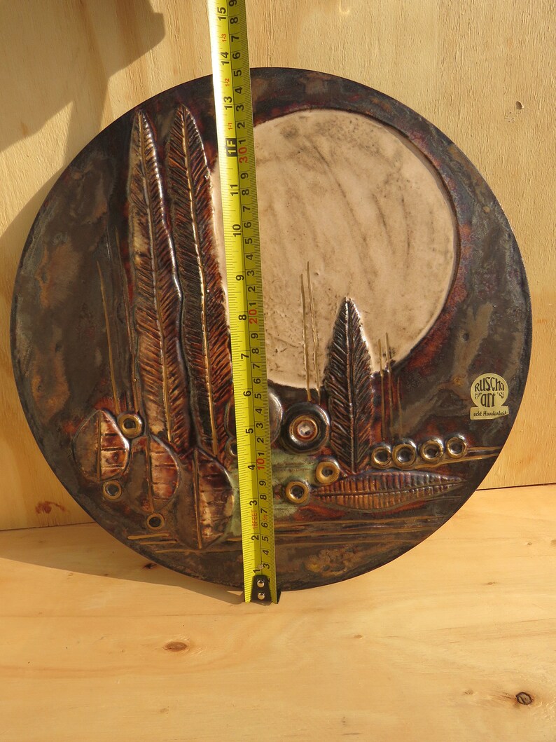Op de afbeelding: Een rond keramisch wandkunstwerk met een bruine, roestkleurige en witte glazuur. De kunst toont een gestileerd ontwerp van bomen en een maan. Het stuk is ongeveer 38 cm in diameter. De kunst is rechtsonder getekend met "Ruscho Art".
