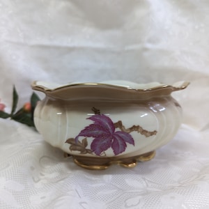 Könnte beinhalten: Ein weißer Keramiktopf mit einem gewellten Rand und goldenen Verzierungen. Der Topf zeigt ein purpurfarbenes und braunes Blumenmuster.