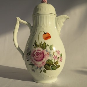 Può includere: Teiera in porcellana bianca con un motivo floreale. La teiera ha un coperchio con un fiore rosa in cima. Il motivo floreale comprende una rosa rosa, un papavero rosso e altri fiori più piccoli in tonalità rosa, bianco e viola.