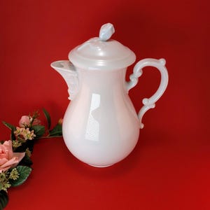Op de afbeelding: Een witte porseleinen koffiepot met een deksel en een gebogen handvat. De pot heeft een parelmoer afwerking en een decoratieve tuit. Een klein bloemaccent staat op het deksel. De achtergrond is effen rood.