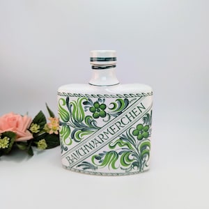May include: White ceramic bottle with a square body and a rounded neck. The bottle is decorated with green floral patterns and the word "BAUCHWÄRMERCHEN" in a diagonal band. The bottle has a white cap with green stripes.