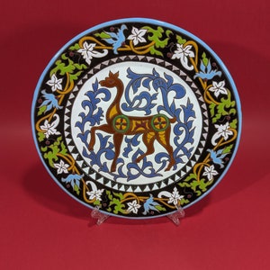 May include: Ceramic decorative plate with a light blue rim. The central design features a brown deer-like animal surrounded by blue and white floral patterns. The plate has a black border with white flowers and green leaves.