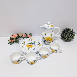 Könnte beinhalten: Ein weißes Keramik-Teeservice mit gelben Blumenmustern. Das Set umfasst eine Teekanne, vier Tassen und mehrere Teller. Jedes Teil hat einen blauen Rand. Die Teekanne hat einen Deckel mit passendem Blumenmuster.
