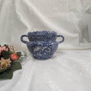 Può includere: Un vaso in ceramica blu e bianco con due manici. Il vaso presenta un motivo dettagliato con un disegno di uccelli e fiori. Il bordo ha un bordo smerlato. Il vaso è posto su uno sfondo bianco a motivi floreali.