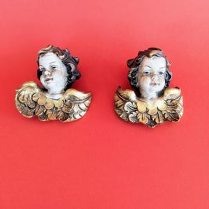 Puede incluir: Dos esculturas de querubines antiguos con rostros detallados y alas doradas. Los querubines tienen piel clara, ojos azules y cabello rizado castaño oscuro. Las alas son de color dorado detallado. El fondo es rojo liso.