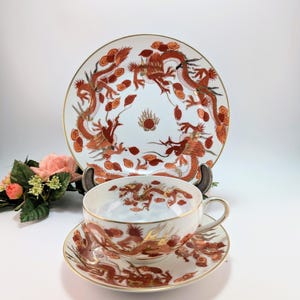Op de afbeelding: Een wit porseleinen theeservies met een rood en goudkleurig drakenontwerp. De set bevat een theekopje, een schotel en een groot bord. De randen van de kop, schotel en bord zijn goudkleurig.