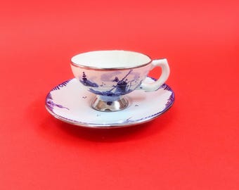 May include: A white teacup and saucer with a blue and white windmill design. The cup has a silver rim and a silver base.