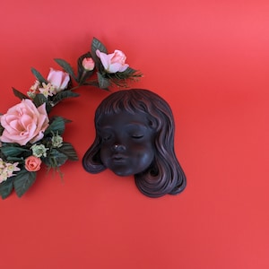 Può includere: Una scultura murale in gesso nero di un viso di donna con gli occhi chiusi e i capelli lunghi. La scultura è posizionata su uno sfondo rosso con un'arrangiamento floreale di rose rosa e foglie verdi.