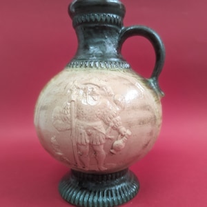 以下が含まれることがあります： 緑色の釉薬がかかった陶器の壺で、鎧を着た男性のレリーフが施されています。壺は丸い胴体と小さな取っ手を持っています。