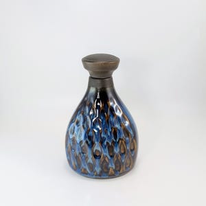 Può includere: Una bottiglia in ceramica con base arrotondata e collo stretto, sormontata da un tappo marrone scuro. Il corpo presenta un motivo testurizzato a smalto colato nei toni del blu profondo, bronzo metallizzato e nero, creando un effetto visivo verticale.
