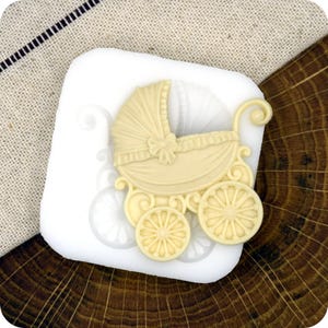 May include: A light yellow baby carriage decoration, featuring a curved hood and spoked wheels, is displayed on a white surface. The carriage has a bow and scrollwork details. The background includes a linen cloth and a wood grain surface.