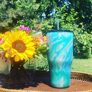 May include: A turquoise and white marbled tumbler with a black lid and a straw. The tumbler is sitting on a brown wicker tray next to a vase of sunflowers and other flowers.
