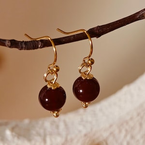 May include: Dangle earrings with deep red, round beads and gold-coloured hooks. The earrings have gold accents at the top and bottom of the beads. They are displayed on a brown branch against a soft, neutral background. The earrings are a jewellery item.