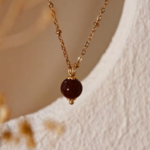 May include: A gold necklace featuring a dark red, spherical pendant. The chain has small, evenly spaced beads. The pendant is a smooth, polished sphere. The necklace hangs against a soft, white backdrop.