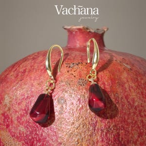 Può includere: Orecchini color oro con un pendente a goccia in pietra preziosa rosso scuro. Gli orecchini sono esposti su un melograno rosso. Il marchio "Vachana jewelry" è nella parte superiore dell'immagine.