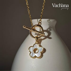 May include: Gold-toned necklace with a flower pendant. The pendant features a white mother-of-pearl flower design with a gold outline. The necklace has a toggle clasp and a delicate chain. The brand name "Vachana jewelry" is visible in the upper right corner.