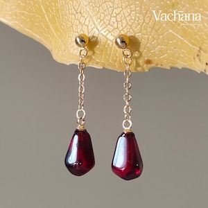 May include: A pair of gold dangle earrings with red teardrop-shaped stones. The earrings are on a yellow leaf background.