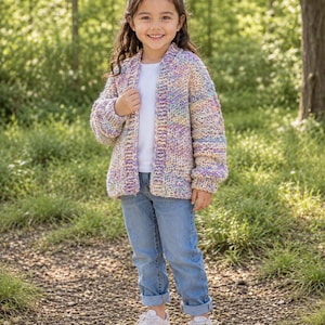 May include: A young person wearing a colorful, chunky knit cardigan with shades of purple, yellow, and blue. The cardigan is paired with a white t-shirt, blue jeans, and white sneakers. The background is a blurred outdoor scene.