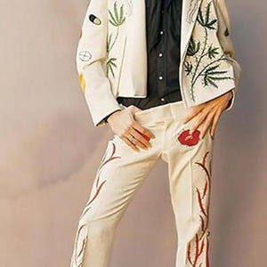 May include: A man wearing a white suit with floral embroidery. The suit jacket has a black shirt underneath. The trousers have red and white floral embroidery. The man is standing on a wooden platform.
