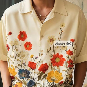 Op de afbeelding: Een crèmekleurig overhemd met korte mouwen en knoopsluiting, voorzien van geborduurde wilde bloemen in oranje, gele en blauwe tinten langs de onderzoom. De tekst Ansari Art staat in het zwart op de rechterkant van de borst.