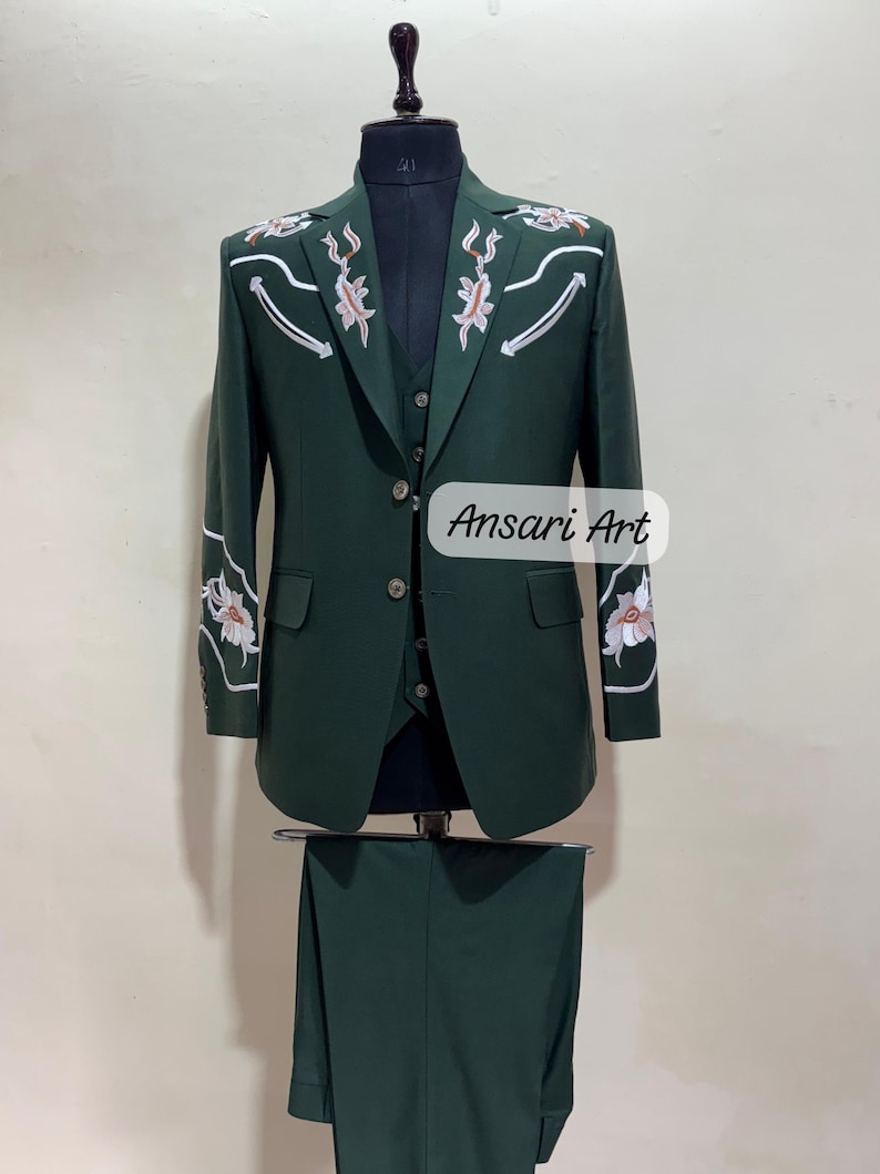May include: A three-piece dark green suit with floral embroidery on the shoulders and sleeves. The suit includes a jacket, vest, and trousers. The text "Ansari Art" is visible on the lower right side of the image.