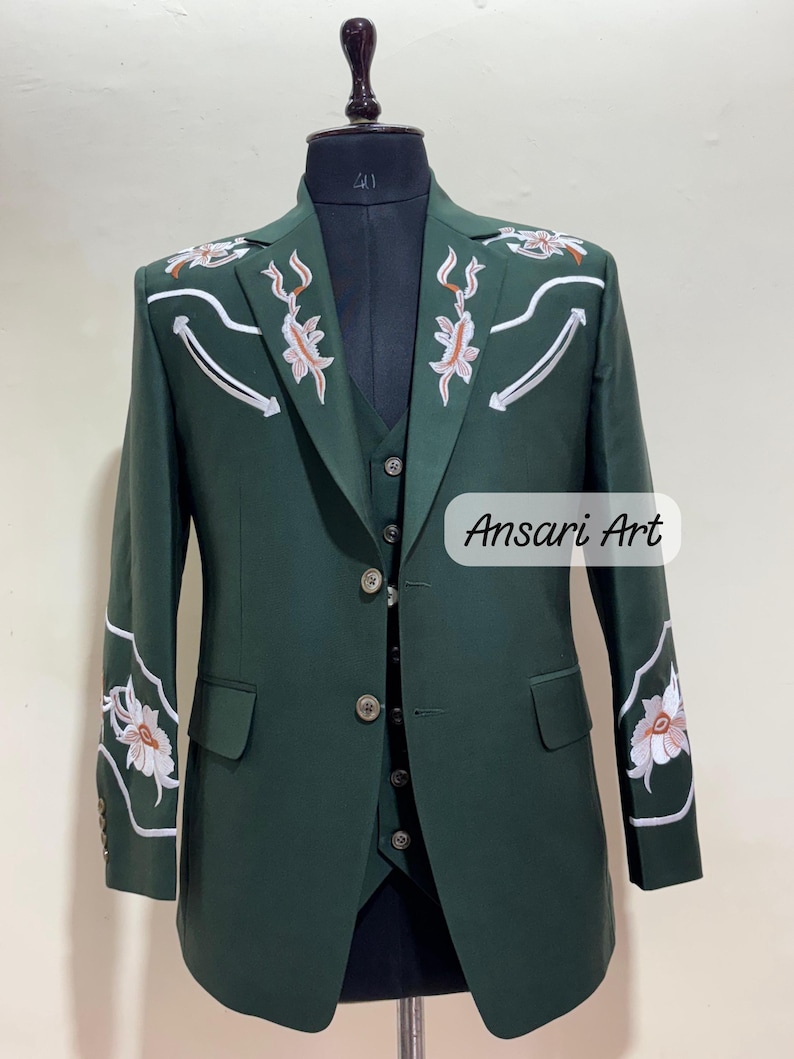 May include: A dark green suit with floral embroidery on the shoulders and sleeves. The suit includes a blazer, vest, and pants, displayed on a mannequin. The text "Ansari Art" is visible on the lower right.