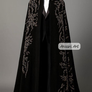 May include: A black cape with ornate gold embroidery, worn over a black suit, white shirt, and bow tie. The cape features a high collar and is displayed on a mannequin. The text "Ansari Art" is visible.