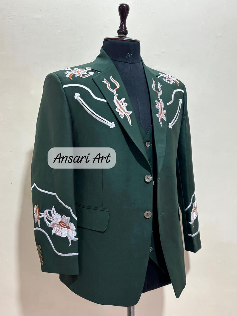 May include: A dark green suit with floral embroidery. The suit includes a blazer and vest, displayed on a mannequin. White and orange floral designs with white accents adorn the shoulders and sleeves. The text "Ansari Art" is visible.