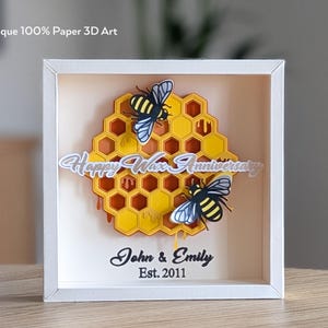 Könnte beinhalten: Ein weiß gerahmtes 3D-Papierkunstwerk. Das Kunstwerk zeigt ein gelb-oranges Wabenmuster mit zwei Bienen. Der Text "Happy Wax Anniversary" befindet sich über der Wabe und "John & Emily Est. 2011" darunter.
